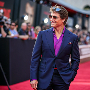 Tom Cruise at the premier of Mission: Impossible. Wearing a sharp navy suit with a vibrant purple shirt underneath, sunglasses perched atop his head. Crowded theater in the background, blurred.