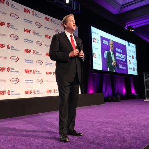Chris Baldwin, CEO of BJ's Wholesale, addressing the NRF Foundation Gala audience. Dressed in a black suit and bright red tie, standing on a deep purple carpet. Behind him, numerous corporate logos are prominently displayed.