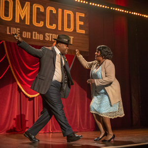 Clark Johnson and Karen Williams, in character as Det. Meldrick Lewis and Barbara Shivers, dancing under spotlight at 'Homicide: Life on the Street' event. Red curtain backdrop with golden banner.