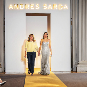 Nuria Sarda and Hiba Abouk on a fashion runway. Hiba in a stunning evening gown, Nuria in casual attire. A spotlight illuminates them as they walk towards the audience. The backdrop features the brand name 'ANDRES SARDA' in large, illuminated letters.