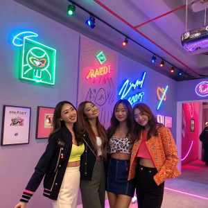 Soyou, Hyorin, Bora, and Dasom of SISTAR at a pop-up shop in Seoul. The room is decorated with neon signs and modern art pieces. They pose for pictures with fans.