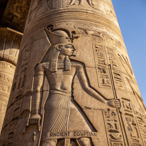 Close-up of an ancient Egyptian column, depicting a powerful pharaoh in intricate detail. The deity stands with authority, adorned with elaborate headdress and regalia. Hieroglyphs cover the weathered stone, showcasing the artistry of ancient Egyptian carvings.