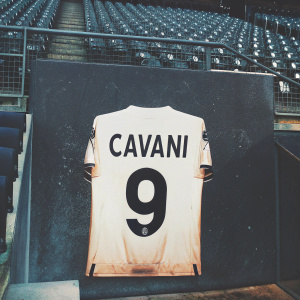 Edinson Cavani's white soccer jersey, classic cut, number 9 and name 'CAVANI' in bold black. Posing against an empty stadium backdrop.