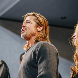 Brad Pitt, sporting a long blonde mane and a rugged beard, captures attention at a Hollywood film premiere. Dressed in a charcoal sweater, he exudes intensity against a moody, dimly lit backdrop.