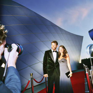 Liam McIntyre and Erin Hasan at a gala event. Smiling, dressed in formal attire. QuA brand backdrop