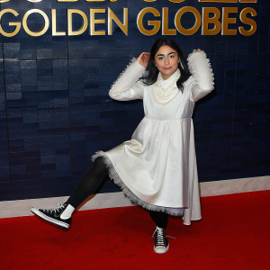 Rosa Salazar at the Golden Globes, standing confidently in a white dress with fluffy trim. She wears black Converse sneakers. Gold text on black backdrop
