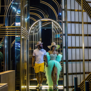 Cardi B and Dan Bilzerian at a glamorous party. Cardi B in an eye-catching light green feathered gown with a wide-brimmed hat. Dan Bilzerian in a white t-shirt, yellow shorts, and a casual beige hat. The backdrop features a luxurious gold and black design.
