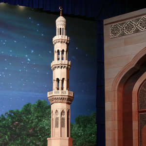 Sultan Qaboos Grand Mosque in Muscat, Oman. Majestic multi-tiered minaret with detailed carvings and ornate stonework. Light brown stone exterior with intricate designs.