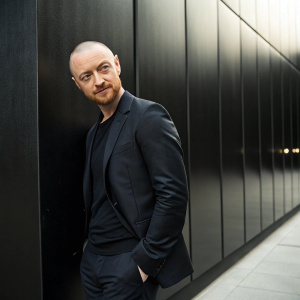 James McAvoy, bald and with stubble, posing for a formal portrait. The background is a sleek black wall.