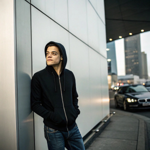 Rami Malek standing in front of a sleek black urban setting. Wearing a black hoodie and jeans. No backdrop of 'Mr. Robot' poster
