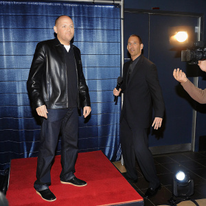Bill Burr and Rich Vos at the Comedy Central Awards. Rich Vos in a black leather jacket, Bill Burr in a dark suit. They are posing on a stage with a red carpet backdrop and flashing cameras.