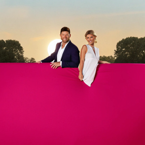 Jensen Ackles and Joanna Krupa, outdoors at sunset, smiling and posing. He's in a navy blazer with a crisp white shirt. She's in a flowing white dress. Lush green trees and a clear sky in background.