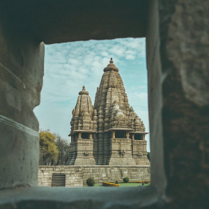 Kandariya Mahadev Temple, an ancient Hindu temple located in India. Multiple levels and towers, constructed from stone with intricate carvings. Set amidst lush greenery, under a clear blue sky dotted with clouds.