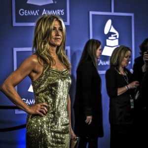 Jennifer Aniston at the Grammy Awards. Wearing a shimmering gold sequined dress. Confident pose. Backdrop features Grammy logo and celebrities around