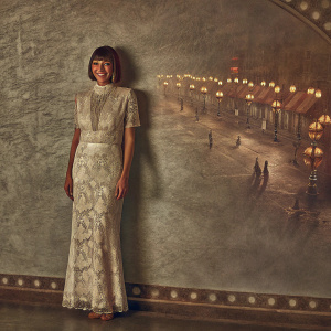 Actress Rashida Jones, with a sleek bob haircut, smiling warmly. Wearing a sophisticated white gown with intricate lace patterns. The backdrop is a bustling street scene at night.
