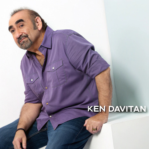 Ken Davitian, actor with a rugged look, in a casual purple button-down shirt. He's at a tech conference posing against a minimalist white backdrop with a sponsor's logo.