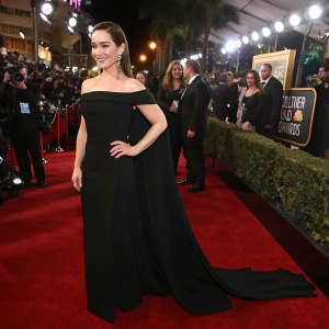 Emilia Clarke at the Golden Globe Awards. Wearing a long black dress with off-the-shoulder design and a flowing train. Crowded event, with flashing cameras and stars around