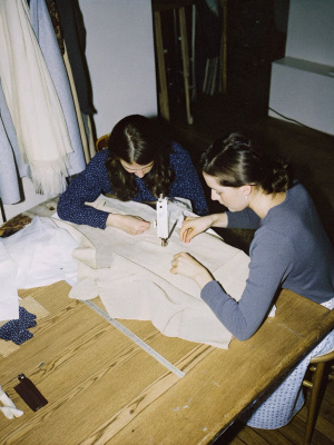 Two seamstresses sewing a minimalist wedding dress with threads, patterns and white fabrics at their side