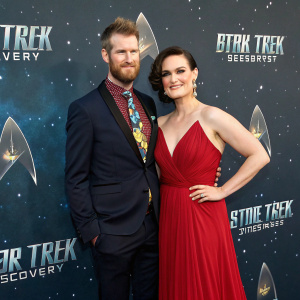 Kenneth Mitchell and Mary Chieffo at the Star Trek Discovery premier. Both dressed formally, Kenneth in a dark suit with a vibrant tie, Mary in a flowing red gown. They pose against a backdrop filled with the iconic Star Trek symbols.