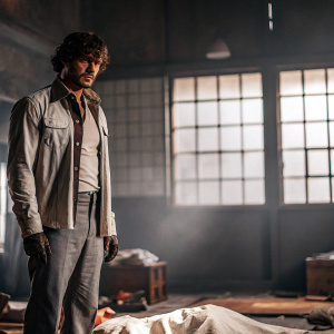 Will Graham, the protagonist from 'Hannibal,' stands amidst a crime scene. Wearing a grim expression, his face is smeared with blood. The dimly lit room with scattered evidence in the background.