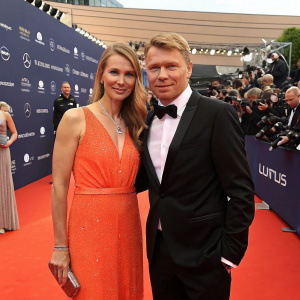 Mika Hakkinen and his wife attending the Laureus World Sports Awards. He is in a sharp black suit. She wears a dazzling orange dress. Crowded red carpet with flashing cameras.