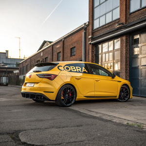 A bright yellow Seat Leon FR Cobra Sport parked in front of a warehouse with Cobra logo. The car's aggressive design, including its sporty exhaust and sleek profile, contrasts against the backdrop of brick walls and building facades illuminated by the afternoon sun.