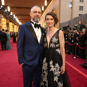 Jason Connery and Mia Sara attending an awards ceremony. Jason in a navy blue tuxedo, Mia in a floor-length black gown with intricate white lace detailing. Crowded venue with red carpet and flashing cameras.
