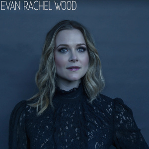 Close-up portrait of actress Evan Rachel Wood. Wearing a black dress with intricate gold embroidery. Soft makeup, wavy blonde hair. Dark studio backdrop with minimal text logo