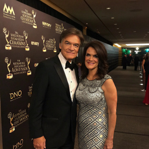 Dr Mehmet Oz and Lisa Oz attending the Daytime Emmy Awards Gala. Dr Oz in a black tuxedo, Lisa in a silver and black patterned dress. The venue has dark walls adorned with various award logos.