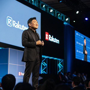 Hiroshi Mikitani, CEO of Rakuten Group Inc., addressing an enthusiastic crowd at a tech summit. He's dressed in a sleek black suit and appears focused. The stage is filled with digital screens displaying the company's logo.
