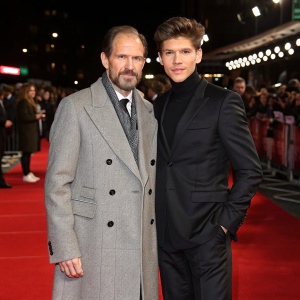 Formal event photo of Ralph Fiennes and Oleg Ivenko at 'The White Crow' premiere. Gray trench coat and black suit. Crowd in background