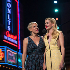 Busy Philipps and Michelle Williams at award show. Dimly lit stage with dramatic spotlight. Neon signs and vibrant backdrop