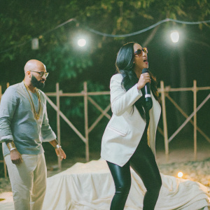 Monica, in a sleek white blazer and black leggings, performs on a brightly lit stage. Jermain Dupri, bald with gold accessories, stands nearby.