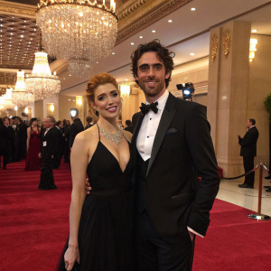 Elena Satine and Tyson Ritter attending a gala event. The venue is opulently decorated with chandeliers and red carpets.