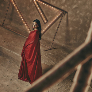 Aishwarya Rai, wearing a deep red sari, poses in a dimly lit studio. Neon signs in the background illuminate the scene. Her dark hair cascades over her shoulders. Close-up shot captures her makeup and radiant face.