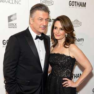 Alec Baldwin and Tina Fey at the Gotham Magazine Gala. They stand gracefully in front of a minimalist white backdrop, adorned with Gotham Magazine's logo. Baldwin, dressed in a sleek black tuxedo, and Fey, wearing a sophisticated black gown with lace details, exude poise and elegance.