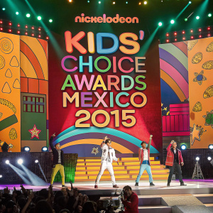 At the lively Nickelodeon Kids' Choice Awards Mexico 2015, a diverse group of performers on a vividly decorated stage. Multi-colored background with large graphics and lettering.