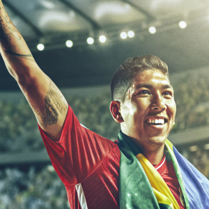 Roberto Firmino, Brazilian soccer star, wearing Liverpool FC jersey. Joyful smile, short hair, tattoo visible. Crowded stadium blurred in background.