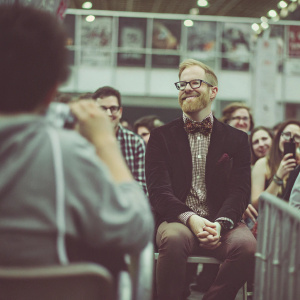 Jesse Tyler Ferguson attending a Q&A session at New York Comic Con. Wearing a dark blazer with a bow tie and patterned shirt. Sitting in front of a crowd of fans.