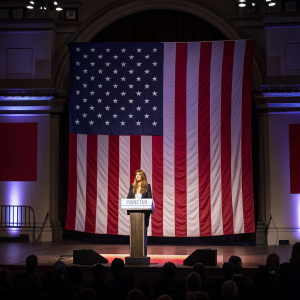 Melania Trump, speaking in front of a large American flag backdrop. The event is taking place in a dimly lit auditorium.
