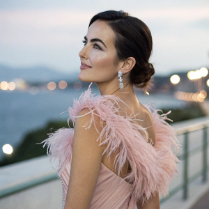 Camilla Belle in an elegant evening gown, adorned with a pink feather accent. Starring at the camera with a subtle smile. Soft, blurred background with lights in the distance.