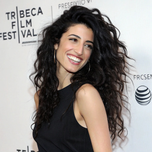 Tania Raymonde, in her late twenties, sporting long curly black hair, grinning brightly. She wears a simple black sleeveless top. Photo taken against a plain white wall with event logos. Event promotional photo style.