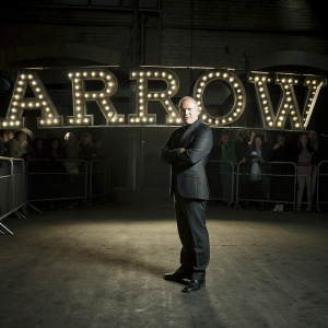 Victor Garber at Comic-Con in a dark suit. He's standing in front of a large, illuminated 'Arrow' banner.