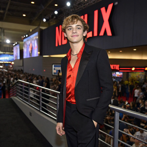 Ross Lynch, actor and singer, at Comic-Con. Wearing black suit with red shirt. Netflix sign in background. Crowded convention floor