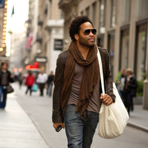 Lenny Kravitz, casually dressed in a brown scarf and sunglasses, walking through a bustling downtown area with a white tote bag.