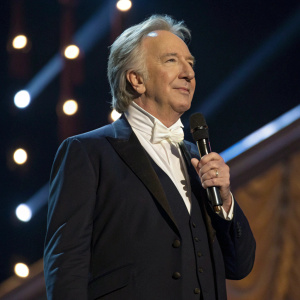 Alan Rickman, British actor, in a dark suit with white cravat. On stage at award show, with microphone in hand.