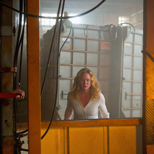 Erika Christensen as Julia Braverman-Graham. She is in a tense scene from the TV series Parenthood. White blouse, wavy blonde hair. Inside a warehouse with industrial equipment, wires, and a large metal container in the background.