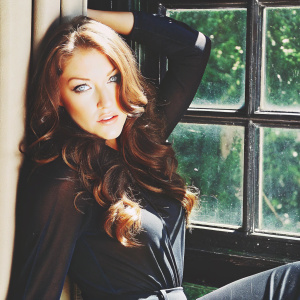 Skyler Samuels, with long brown wavy hair and blue eyes, posing for a studio portrait. Soft natural lighting, green backdrop