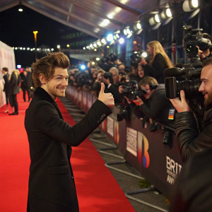 Celebrity giving a thumbs-up at the Brit Awards. Crowded red carpet with flashing cameras.