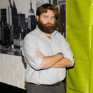 Zach Galifianakis in a candid shot. Serious face, thick beard, blue shirt. Cityscape background
